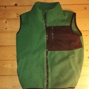 Grön fleeceväst med svart ficka - Grön fleeceväst med hög krage och svart bröstficka i nylonmaterial med dragkedja. Västen har hel dragkedja framtill och svarta kantband vid ärmöppningarna. Perfekt för lager-på-lager och chill utomhus.