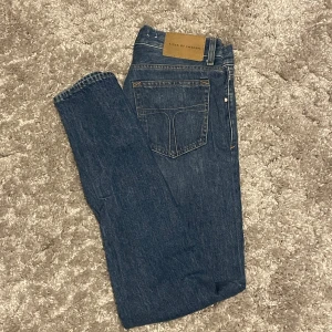 Blå jeans från Tiger of Sweden - Snygga blå jeans från Tiger of Sweden med klassisk femficksdesign och läderpatch bak i midjan. Jeansen har raka ben och är tillverkade i robust denim som ger en tidlös look. Perfekta för dig som gillar stilrena och enkla jeans.