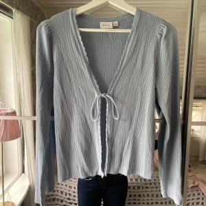 Ljusblå kofta  - Ljusblå ribbad kofta från Vila med lång ärm och v-ringning. Koftan har volangkant längs ringningen och knyt framtill. 