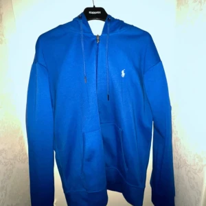 Polo Ralph Lauren Zip Hoodie - Snygg blå hoodie från Ralph Lauren med klassisk vit logga på bröstet. Tröjan har huva med snörning, magficka och långa ärmar. Perfekt för dig som gillar en stilren och sportig look.
