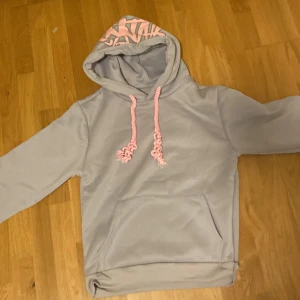 Ljusgrå hoodie med rosa detaljer - Säljer en ljusgrå hoodie med känguruficka och huva. Huvan har ett coolt rosa zebramönster på insidan och matchande rosa snören. Perfekt för dig som gillar en chill och snygg stil.