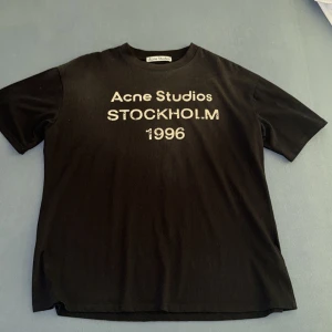 Svart Acne Studios t-shirt Stockholm - Svart t-shirt från Acne Studios med tryck 'Acne Studios STOCKHOLM 1996' på bröstet. Klassisk passform med rund hals och korta ärmar. Tillverkad i 100% ekologisk bomull för en skön och mjuk känsla. Perfekt för en clean och stilren look.