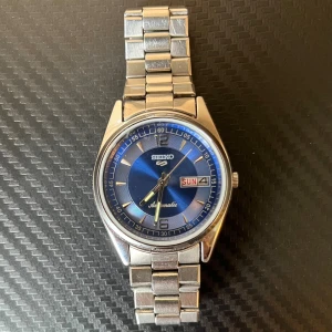 Seiko 5 - Snygg Seiko 5 automatisk klocka med blå urtavla och dag/datumvisning. Klockan har ett klassiskt runt boett i silverfärgat stål och matchande länkarmband. Mekaniskt urverk syns genom baksidan. Perfekt för dig som gillar stilren design och japansk kvalitet.