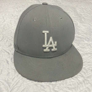 Grå LA Dodgers keps från New Era - Säljer en stilren grå New Era 59FIFTY keps med LA Dodgers-logga broderad framtill i vitt. Klassisk modell med böjd skärm och MLB-logga baktill. Perfekt för dig som gillar sportig streetstyle och vill ha en clean look.