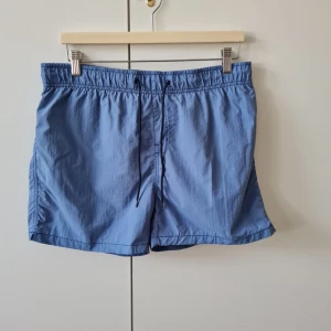Badbyxor med snörning från Cubus strl L - Snygga blå badshorts från Cubus i storlek L (känns som storlek M). De har elastisk midja med svart snörning och en bakficka med knapp. Tillverkade i lätt syntetmaterial med meshfoder, perfekta för strand eller pool. Kan hämtas i Vallentuna.