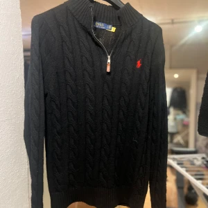 Svart kabelstickad tröja Polo Ralph Lauren - Svart kabelstickad tröja från Polo Ralph Lauren med halv dragkedja och röd broderad logga på bröstet. Tröjan har hög krage, långa ärmar och ribbade muddar. Perfekt för dig som gillar klassisk och stilren look.