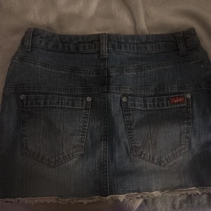 Jeanskjol med spetskant  - Mörkblå jeanskjol som är egen sydd med klassiska bakfickor, bälteshällor och en snygg spetskant längst ner. Kjolen har en rak passform och är i kort modell, perfekt för dig som gillar denim med en söt twist.🥰 passar dig med 34/36. S eller xs! Pris kan diskuteras vid snabb affär