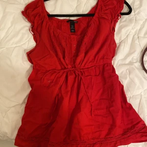 Röd blus med spets och knyt i midjan - Snygg röd blus från H&M med spetsdetaljer framtill, knappar vid bysten och knytband i midjan. Blusen har korta volangärmar och en luftig passform, perfekt för varmare dagar eller nu till hösten. Tillverkad i mjuk bomull för en skön känsla.
