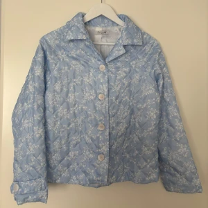 Blå/vit jacka  - ljusblå quiltad jacka med vitt blommönster från Design by Si. Köpt secondhand/ sellpy, inte kommit till användning. 