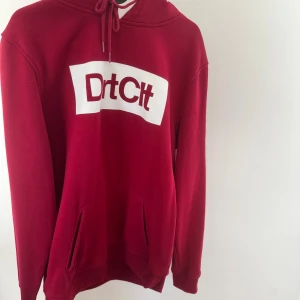 Röd hoodie från DirtCült  - Säljer en röd hoodie från DirtCült med vit logga framtill. Tröjan har huva med snörning, känguruficka och långa ärmar. Perfekt för dig som gillar streetwear och vill ha en skön och snygg look.