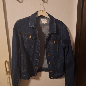 Mörkblå jeansjacka från Vila - Snygg mörkblå jeansjacka från Vila med klassisk passform. Jackan har knappar i guldton, två bröstfickor med lock, sidofickorna är inte riktiga. Perfekt för lager-på-lager och enkel att styla till olika outfits.