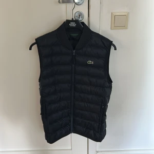 Svart dunväst från Lacoste - Svart dunväst från Lacoste med klassisk quiltad design och dragkedja framtill. Västen har ribbad krage och ikoniska krokodilloggan på bröstet. Perfekt för lager-på-lager och snygg till både hoodie och t-shirt.