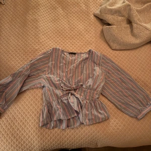 Randig blus från Zara - Säljer en luftig blus med blå bas och tunna ränder i vitt och rött. Blusen har omlottmodell med knyt framtill, v-ringning och långa ärmar med lätt volang nertill. Perfekt för dig som gillar en chill men ändå snygg stil.
