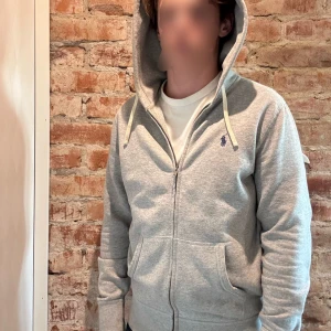Grå hoodie från Polo Ralph Lauren - Snygg grå hoodie från Polo Ralph Lauren med dragkedja, huva med snören och klassisk blå logga på bröstet. Mjuk bomullskänsla och känguruficka framtill. Perfekt nu för hösten/vintern när de börjar bli kyligt!🤩