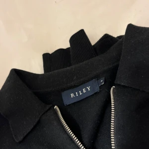 Svart ziptröja från Riley - Svart långärmad tröja från Riley med krage och hel dragkedja framtill. Tröjan har en clean look och är tillverkad i ett mjukt material som känns skönt mot huden. Perfekt för dig som gillar stilrena och enkla plagg.