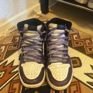 Nike Air Jordan 1 Court Purple High - Säljer ett par Nike Air Jordan 1 High i färgen Court Purple. Skorna har en snygg mix av vitt och lila skinn med svarta detaljer och klassisk svart swoosh. Lila snören och hög siluett ger en riktigt clean och sportig vibe. Perfekta för dig som vill sticka ut med dina sneakers.