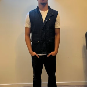 Svart quiltad dunväst från Bläck - Svart quiltad dunväst från Bläck med ståkrage och tryckknappar framtill. Västen har två stora fickor med lock och en bröstficka med dragkedja. Snygg och enkel design som passar till många outfits.