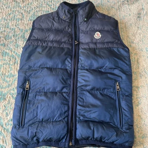 Blå dunväst från Moncler - Snygg marinblå dunväst från Moncler med klassisk logga på bröstet. Västen har hög krage, två fickor med dragkedja och quiltad design. Perfekt för lager-på-lager och ger en sportig vibe. Innerfoder med serietidningsmönster och autenticitetsmärkning.