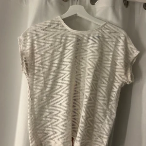 Vit mönstrad blus med brunt band - Säljer en vit blus med transparent zickzack-mönster och korta ärmar. På baksidan finns ett brunt dekorativt band med små metallnitar som ger en cool kontrast. Perfekt för dig som gillar unika detaljer och luftig stil.