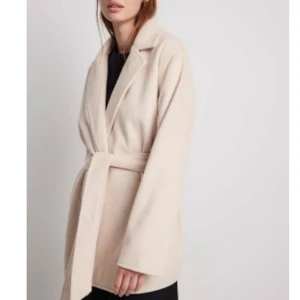 Beige omlottkappa från Ida Carlsson x NA-KD - Snygg beige kappa från Ida Carlsson x NA-KD med omlottmodell och bälte i midjan. Jackan har också fickor på sidan. Helt ny, endast använd 1 gång.. Köpt för 899 kr, säljer för 369 kr. Storlek 32 men stor i storlek. 
