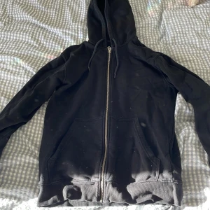 Svart hoodie med dragkedja från Weekday - Svart hoodie från Weekday i storlek XS. Klassisk modell med huva, snörning och dragkedja framtill. Mjukt bomullsmaterial och två stora fickor framtill. Perfekt för en chill och avslappnad stil.