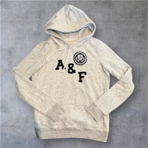Ljusgrå hoodie från Abercrombie & Fitch - Snygg ljusgrå hoodie från Abercrombie & Fitch med broderad A&F-logga och emblem på bröstet. Tröjan har huva med snörning, långa ärmar och en klassisk känguruficka framtill. Perfekt för en chill och avslappnad stil.