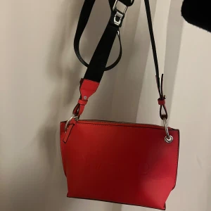 Röd axelväska från NA-KD i skinnimitation - Snygg röd axelväska från NA-KD med bred svart axelrem med logga. Väskan är i skinnimitation och har silverfärgade metalldetaljer samt dragkedja upptill. Justerbar axelrem. 