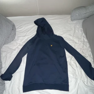 Mörkblå hoodie från Lyle & Scott - Mörkblå hoodie från Lyle & Scott med klassisk gul logga på bröstet. Tröjan har huva, känguruficka och långa ärmar. Perfekt för dig som gillar stilren och enkel design. Materialet är mjukt och skönt, troligtvis bomull eller bomullsblandning.