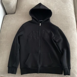 Tuttolente MT Triple Black Hoodie - Svart hoodie med dragkedja och huva. Framsidan har broderade bokstäver och baksidan pryds av en stor svart örn. Tröjan har långa ärmar, ribbade muddar och två fickor framtill. Perfekt för dig som gillar streetwear med attityd.