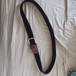 Svart flätat bälte med skinn-detaljer - Snyggt svart flätat bälte med bruna skinn-detaljer och silvrigt spänne. Bältet har en klassisk look med kontrastsömmar på skinnet och passar perfekt till jeans eller chinos. Enkel att justera tack vare det flexibla materialet.