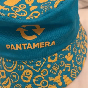 Pantamera bucket hat i blått och gult - Cool bucket hat från Pantamera i klarblå färg med gula tryck och logga. Hatten har massor av roliga, tecknade symboler och emojis över hela ytan. Perfekt för dig som vill sticka ut och visa din hållbarhetsstil. Tillverkad i bomull. Den är också helt ny