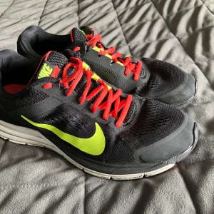 Nike sneakers löparsko - Svarta Nike sneakers med neongul swoosh, röda skosnören och röda detaljer på sulan. Ovandelen är i mesh och syntet för bra andningsförmåga. Perfekta för dig som gillar sportig stil och vill sticka ut med färgglada detaljer.