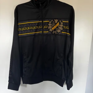 Svart AIK track jacket med brodyr - Svart track jacket från AIK med broderad klubbsköld på bröstet och gul-svarta detaljer över bröstet. Jackan har dragkedja framtill, ståkrage och ribbade muddar. Perfekt för dig som vill visa ditt AIK-stöd med stil.