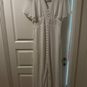 Vit långklänning med knappar och slits - Elegant vit långklänning med v-ringning, korta transparenta ärmar och dekorativa knappar längs hela framsidan. Klänningen har en snygg slits framtill och är tillverkad i ett lätt, flowy material som ger en romantisk känsla.  Jag har bara användt klänningen en gång och det var då jag tog studenten, en jätte fin klänning