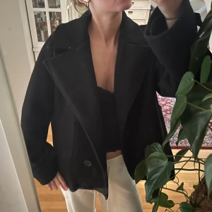 Svart trenchcoat - Säljer en svart trenchcoat med breda slag och stora knappar framtill, ena armen har tappat en knapp (se bild 2). Jackan har långa ärmar med dekorativa knappar vid ärmsluten och en rak, lite oversized passform. Perfekt för dig som gillar klassisk stil med en modern twist. Bra skick