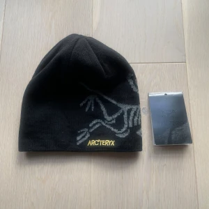 Svart mössa från Arc'teryx - Svart mössa från Arc'teryx med grått logomönster och gul broderad logotyp framtill. Tillverkad i mjukt material som passar perfekt för kyliga dagar. Snygg och stilren design som funkar till de flesta outfits.