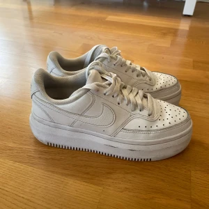 Nike Air Force 1 vita sneakers - Säljer ett par klassiska Nike Air Force 1 sneakers i helvitt skinn med perforerad tå, rund tå och platt sula. Skorna har snörning och den ikoniska swoosh-loggan på sidorna. Perfekta för dig som gillar en clean och tidlös look.