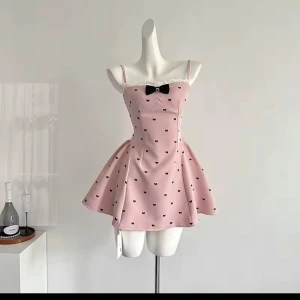 Rosa kortklänning med rosett och spets - Supersöt rosa kortklänning med svarta små rosett-mönster, tunn spetskant upptill och en svart rosett i mitten av bröstet. Klänningen har smala axelband och en utställd kjol som ger en gullig siluett. Perfekt för dig som gillar en feminin och lekfull stil.