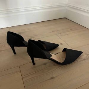 Svarta pumps med spetsig tå från Mango - Snygga svarta pumps från Mango med spetsig tå och låg klack. Skorna har en stilren design med öppen insida och är tillverkade i ett tygmaterial som ger en elegant känsla. Perfekta för dig som vill ha en klassisk och trendig look.