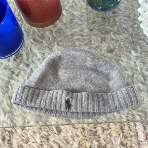 Grå stickad mössa från Polo Ralph Lauren - Snygg grå stickad mössa från Polo Ralph Lauren med klassisk ribbad kant och broderad svart logga framtill.