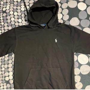 Svart hoodie Ralph Lauren - Svart hoodie från Polo Ralph Lauren med klassisk broderad logga på bröstet. Tröjan har huva med dragsko och en stor magficka framtill. Perfekt för dig som gillar stilren och enkel streetwear.