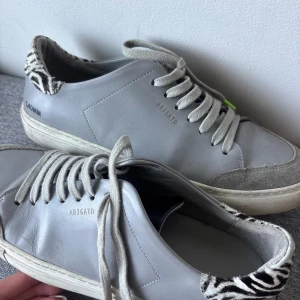 Grå sneakers Axel Arigato zebra-detalj - Grå sneakers från Axel Arigato i skinn med coola zebramönstrade detaljer på hälen. Klassisk låg modell med snörning. Mycket andvända 