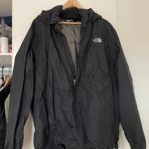 Svart vindjacka från The North Face XL - Svart vindjacka från The North Face i storlek XL. Jackan har huva, dragkedja framtill och meshfoder på insidan. Klassisk logga broderad på bröstet. Perfekt för blåsiga dagar och enkel att matcha med allt.