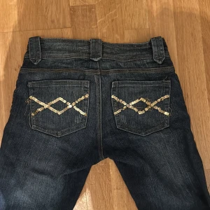 Bootcut jeans med paljettdetaljer - Mörkblå bootcut jeans från Highway med snygga guldpaljetter i mönster på bakfickorna. Jeansen har låg midja, dubbla knappar och klassisk femficksdesign. Dessa är aaas snygga men passar tyvärr inte mig längre. Oklart vilken storlek men skulle säga xs/34 och ungefär 25-26 i midjan.
