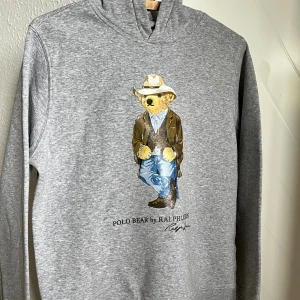 Grå hoodie Polo Bear Ralph Lauren - Grå hoodie från Polo Ralph Lauren med ikoniska Polo Bear-trycket på bröstet. Tröjan har huva och långa ärmar, perfekt för en chill och avslappnad stil. Tillverkad i mjuk bomullsmix för extra komfort.