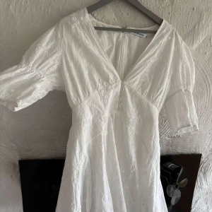 Klänning adoore - Superfin vit klänning från Adoore med broderade blommor, v-ringning och knappar framtill. Klänningen har puffiga korta ärmar och är tillverkad i bomull. Perfekt för sommarens alla tillfällen!