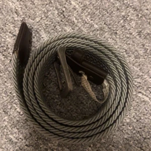 Grått flätat bälte med läderdetaljer - Säljer ett grått flätat bälte med snygga läderdetaljer vid ändarna och metallspänne. Bältet har en stilren och enkel design som passar till många olika outfits. Perfekt för dig som vill ha ett bälte som sticker ut lite men ändå är diskret.