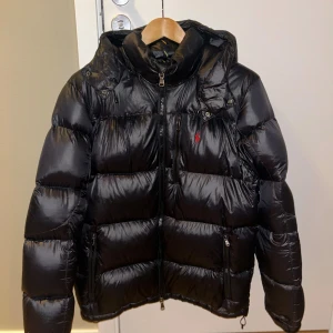Polo Ralph Lauren Puffer Jacket - Snygg svart pufferjacka från Polo Ralph Lauren med glansig finish och röd logga på bröstet. Inga defekter eller problem då den är sparsamt använd och bara använd 2 vintrar. Bara skriva om det är några frågor eller funderingar!