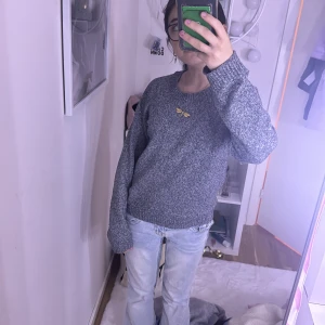 Grå stickad tröja med broderad detalj - Mysig grå stickad tröja från vero Moda med rund halsringning och en broderad gul slända på bröstet. Tröjan har långa ärmar och ribbade muddar vid ärmslut och nederkant. Perfekt för lager-på-lager under kyliga dagar. 