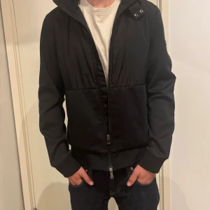 Svart jacka från J.Lindeberg - Snygg svart jacka från J.Lindeberg med diskret logga på ärmen. Jackan har dragkedja framtill, ribbade muddar och en stilren look. Materialet är polyester och jackan har en klassisk passform med lång ärm. Perfekt för dig som gillar en clean och modern stil. Han på bilden är 1,72.